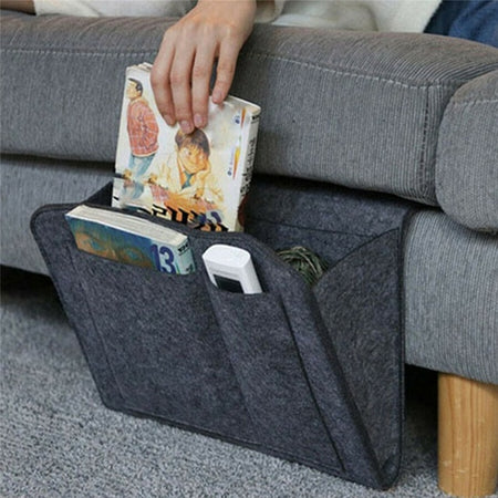 Een grijze BedCaddy met twee boeken en afstandsbediening hangt aan de zijkant van een grijze bank.